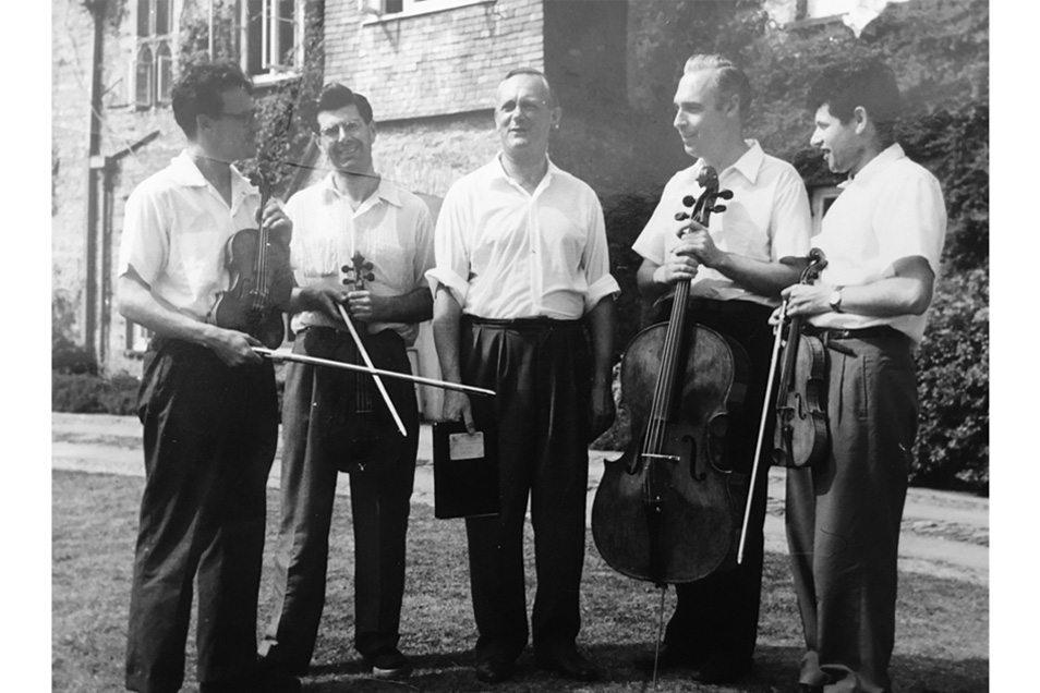 A black and white photo of a group of people wearing shirts and black trousers, carrying string instruments in front of an old brick building.